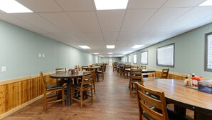 A spacious dining area with multiple tables and chairs, wooden paneling on the walls, and a high ceiling with fluorescent lighting. - Target Hospitality - Big Spring Lodge (Big Spring)