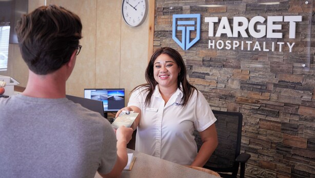 A reception area with a stone wall and a clock. A person is handing a card to a staff member. - Target Hospitality - Big Spring Lodge (Big Spring)
