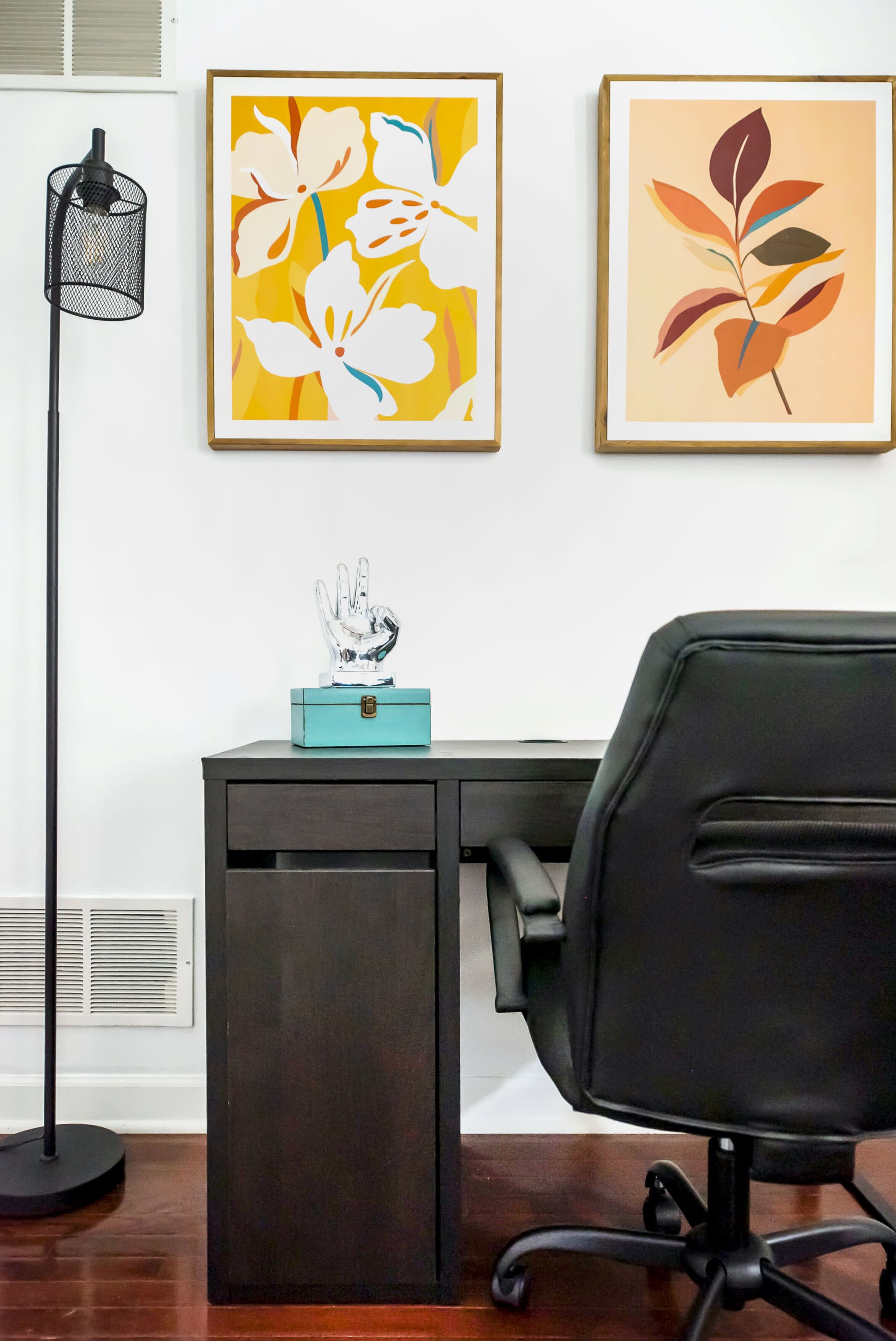 A modern office space with a dark desk, a black office chair, and two framed floral artworks on the wall.