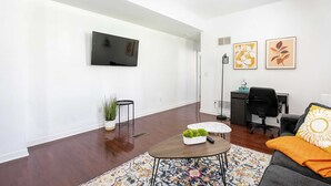 Una sala moderna con televisor de pantalla plana, dos cuadros con motivos florales, un escritorio con silla, y un sofá con una manta.