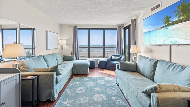 Un salon moderne doté d’une grande fenêtre donnant sur l’océan, d’un tapis bleu aux motifs marins et d’un tableau représentant une scène de plage accroché au mur.