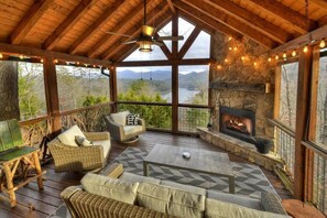 Una terraza de madera acogedora con muebles de mimbre, una chimenea de piedra y una vista panorámica de un río y montañas.