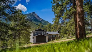 Une maison à deux étages avec un garage, entourée d’une végétation luxuriante et avec une montagne en arrière-plan.