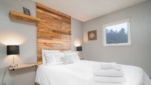 Une chambre à coucher avec une tête de lit en bois, un lit recouvert d’une literie blanche, deux lampes de chevet, une fenêtre donnant sur des arbres et une petite étagère sur laquelle est posé un panneau.