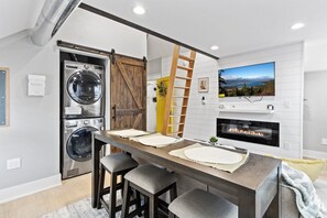 A modern kitchen with a wooden table, bar stools, a built-in washer and dryer, a fireplace, and a TV mounted on the wall. - Live in Chic and Comfort: Experience Our Thoughtfully Designed 1 Bed with Loft! (Germantown)