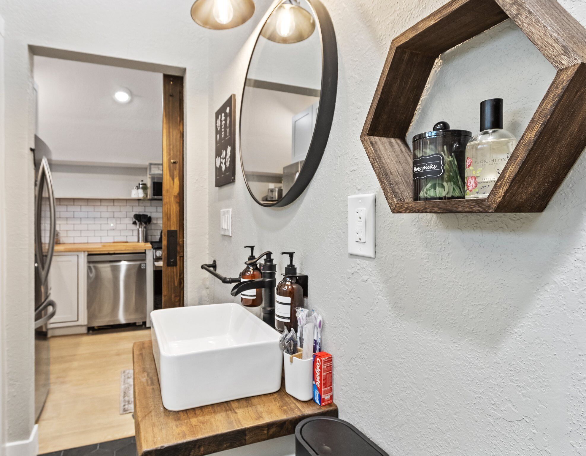 A modern bathroom with a white sink, wooden countertop, and a mirror with a wooden frame. There are toiletries on the counter and a shower area visible in the background.