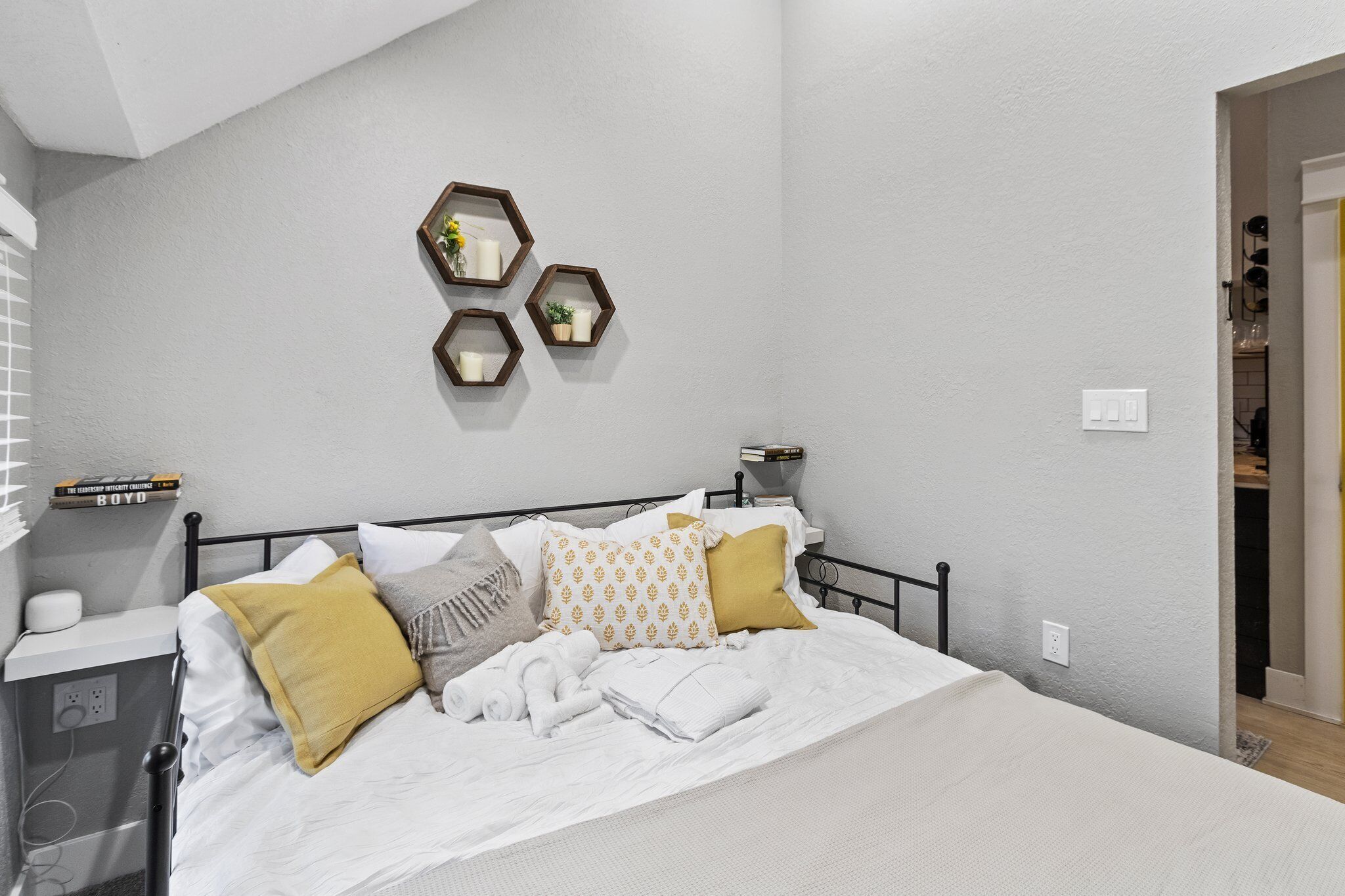 A neatly made bed with white linens and decorative pillows, a bedside table with a lamp, and a wall-mounted shelf with books and a small plant.