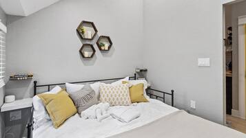 A neatly made bed with white linens and decorative pillows, a bedside table with a lamp, and a wall-mounted shelf with books and a small plant.