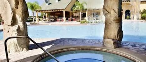 A hot tub under a stone structure with a pool and a building in the background.