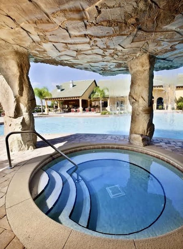 A hot tub under a stone structure with a pool and a building in the background.
