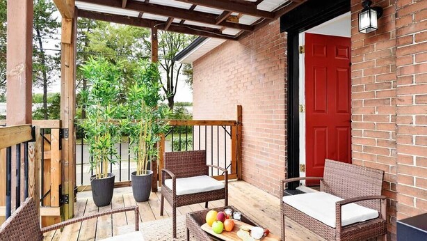 Une terrasse couverte avec des meubles en osier, une table en bois sur laquelle sont disposés des fruits et du pain, et une porte rouge.