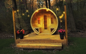 Una cabaña de madera con una entrada circular, rodeada por un bosque. Hay escalones que conducen a la puerta, y dos macetas con flores rojas a cada lado.