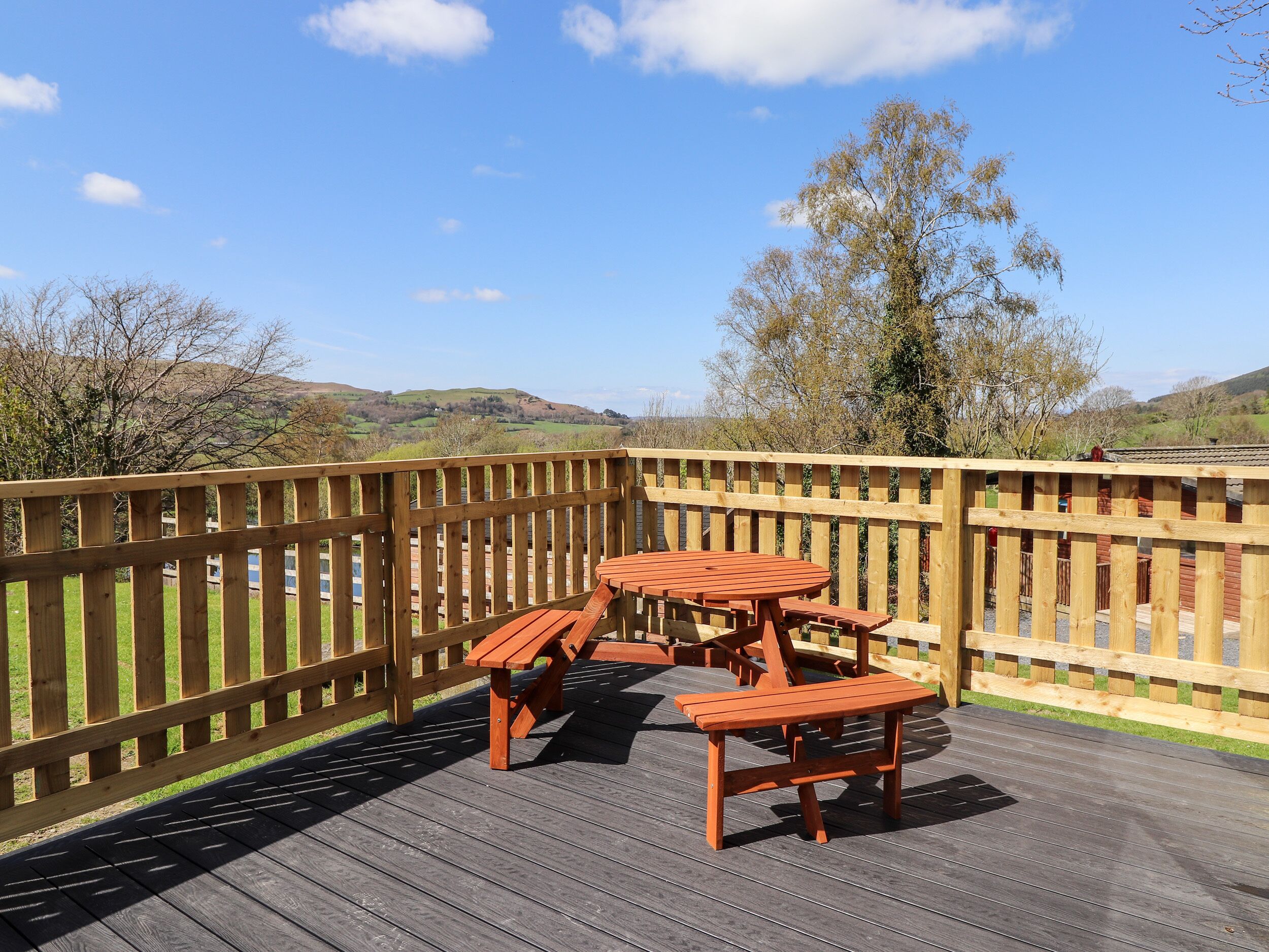 A wooden deck with a picnic table and benches, surrounded by a wooden fence, overlooking a hilly landscape.