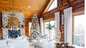 Interior acogedor de una cabaña de troncos con chimenea de piedra, un árbol de Navidad, un sofá blanco y una mesa de comedor de madera.