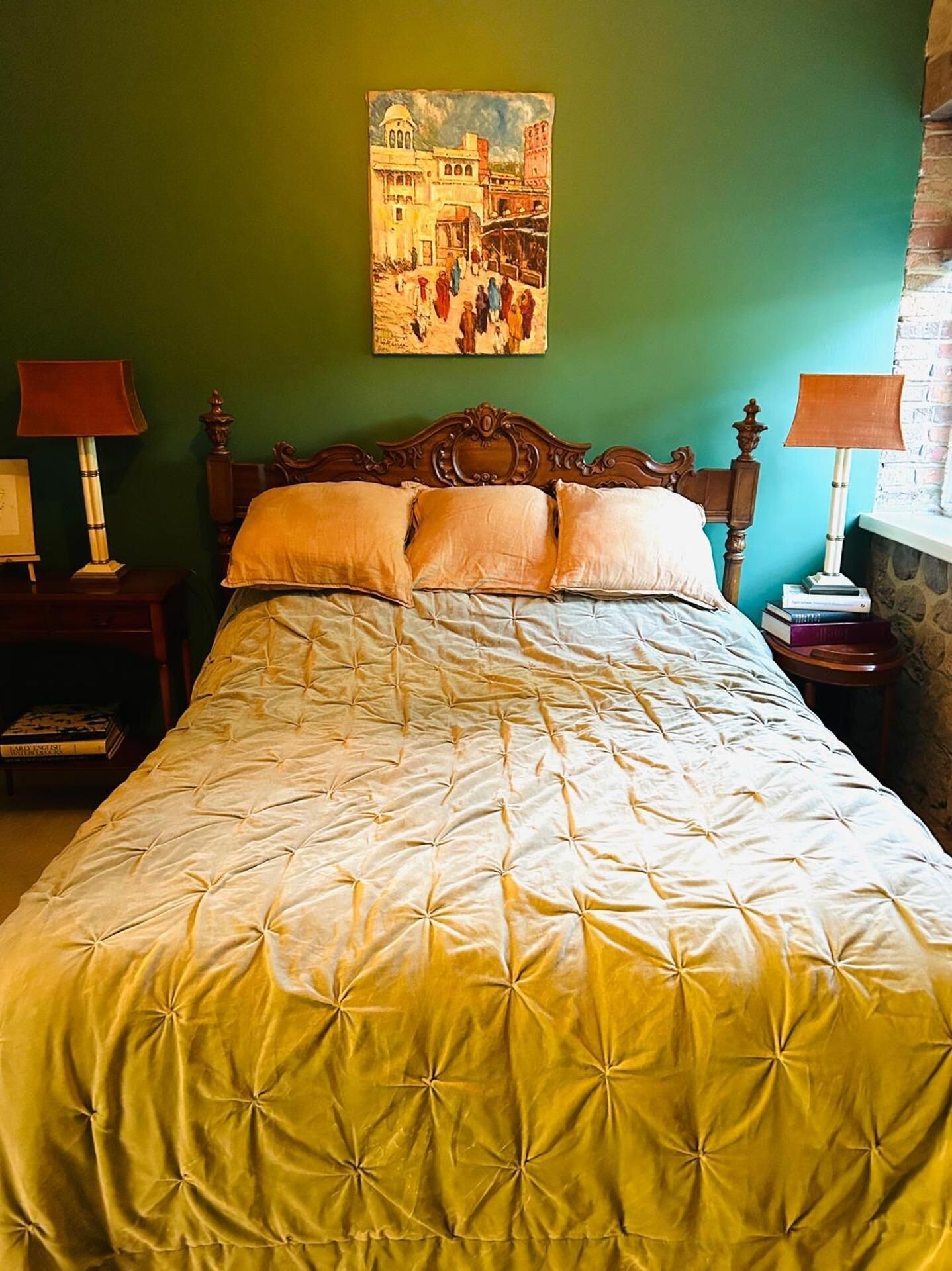A bedroom with a bed, two bedside tables, a lamp, a painting on the wall, and a window with a stone wall.
