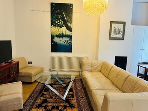 A living room with a sofa, a glass coffee table, a patterned rug, a painting, a radiator, and a chandelier. - No Longer Available (Petworth)