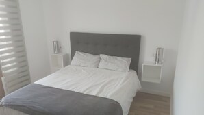 Une chambre moderne avec un grand lit, une literie blanche et une tête de lit grise. On y trouve deux tables de chevet avec des lampes.