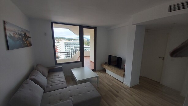 Un salon moderne avec un canapé d’angle, un téléviseur à écran plat posé sur une étagère en bois et une grande porte-fenêtre coulissante donnant sur un balcon avec vue sur la ville.