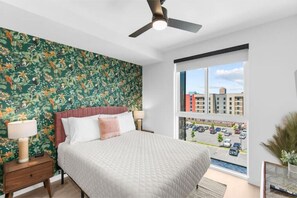 Une chambre avec un grand lit, un ventilateur de plafond, une fenêtre donnant sur un parking et des immeubles, et un papier peint à motifs floraux sur un des murs.