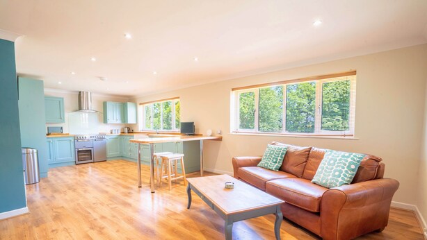A spacious living area with a wooden floor, a brown leather sofa with patterned cushions, a coffee table, and a kitchen in the background. - Long Lane Stables Lodge. Spacious Country Retreat (Penselwood)
