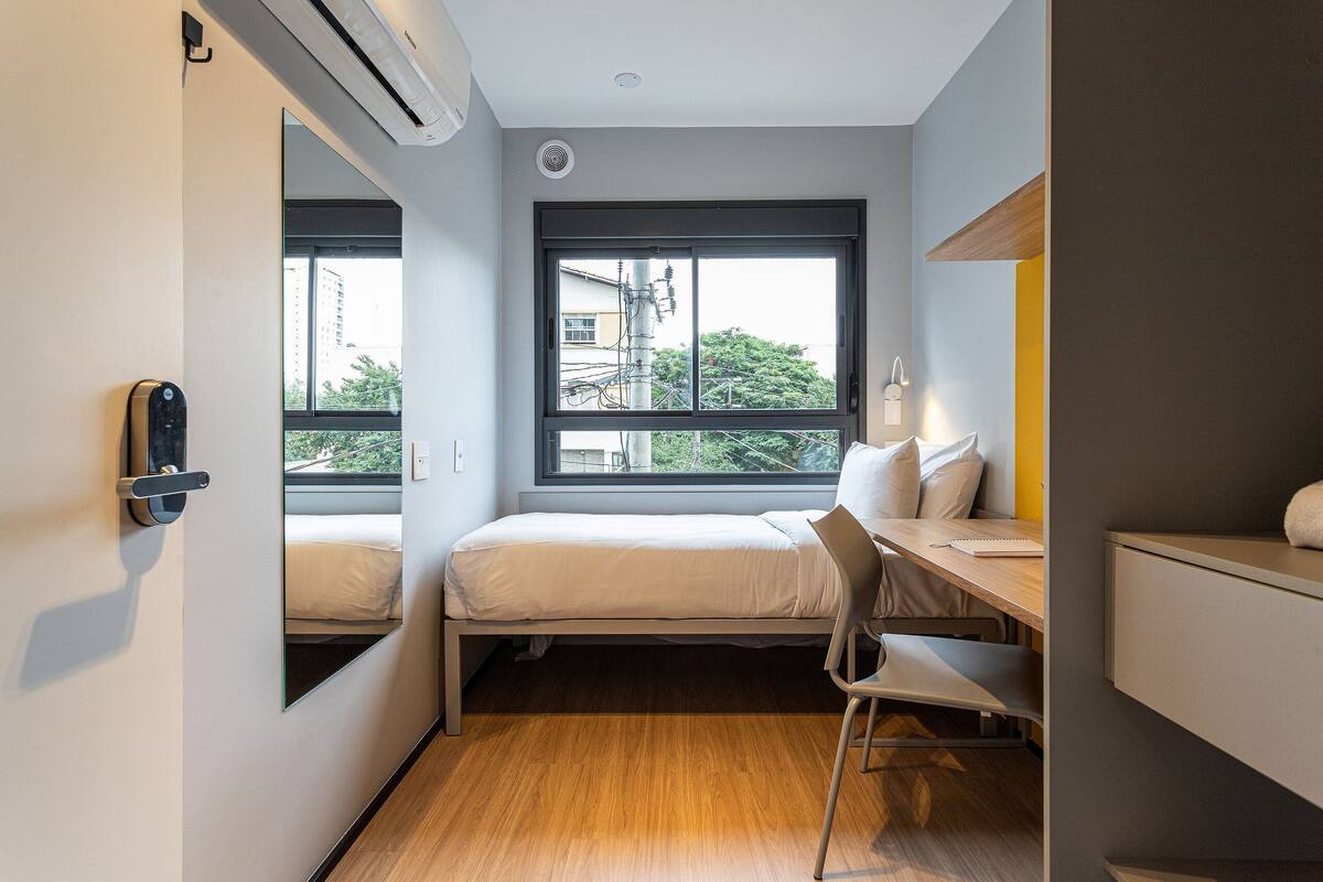 Ein kleines Hotelzimmer mit Bett, Schreibtisch, Stuhl und großem Fenster mit Blick auf die Stadt.