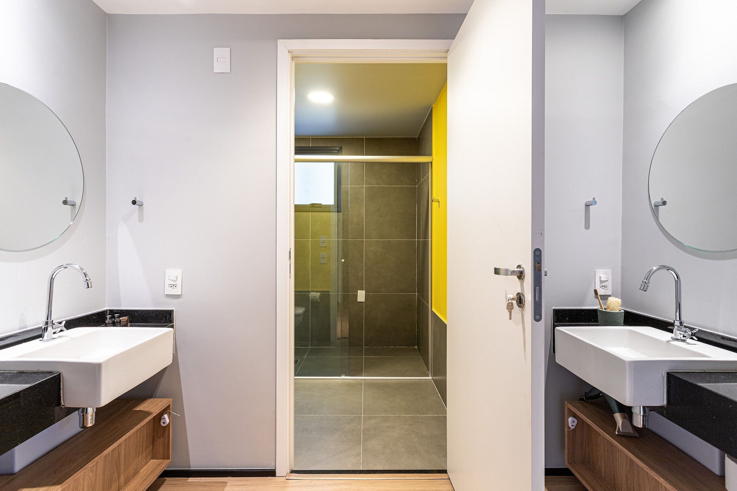 A modern bathroom with two sinks, a shower, and a door leading to a bathroom with a yellow accent wall.