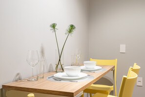 Una mesa de comedor con platos y tazones blancos, dos vasos transparentes y un jarrón con tallos verdes.