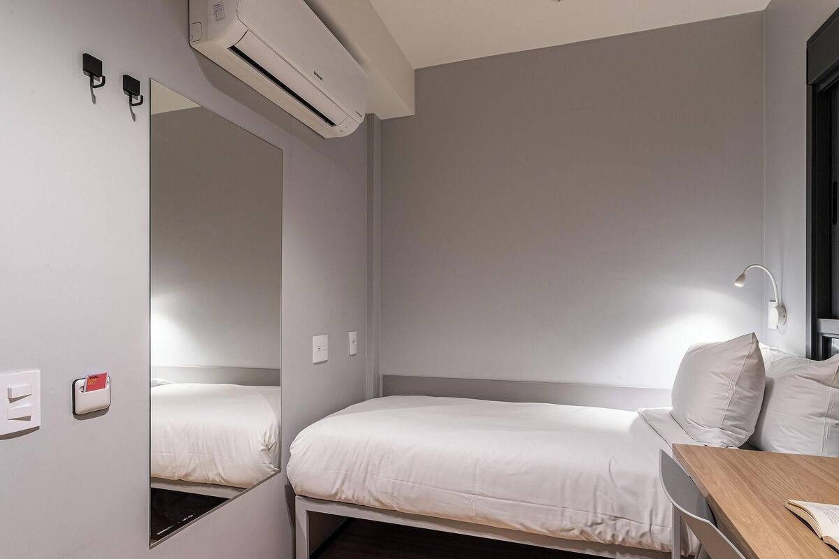 Ein Hotelzimmer mit zwei Einzelbetten, einem Schreibtisch mit Stuhl, einem Fenster mit Jalousien, einer Klimaanlage und einer Wandleuchte.