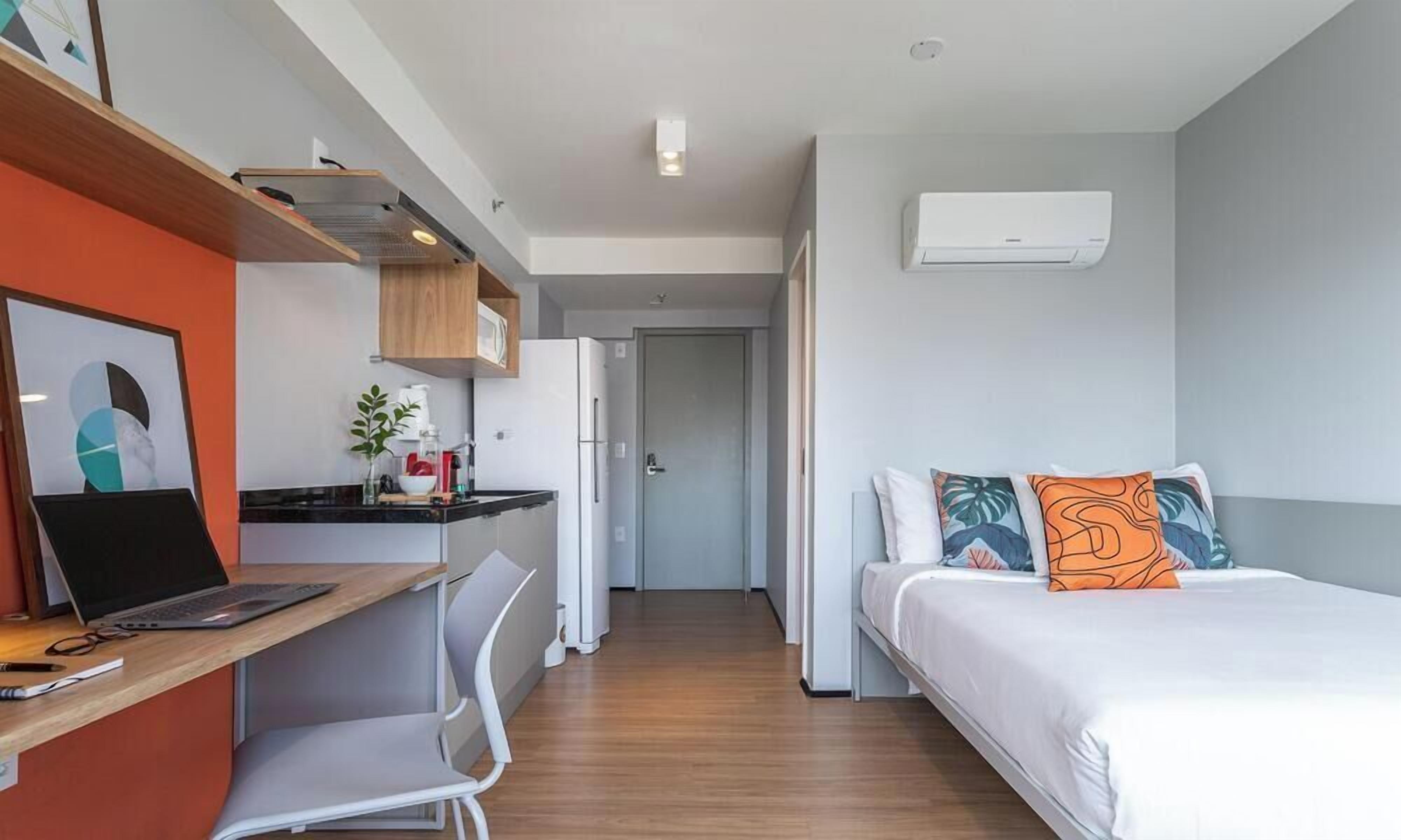 A modern hotel room with a desk, chair, bed, and an air conditioning unit.