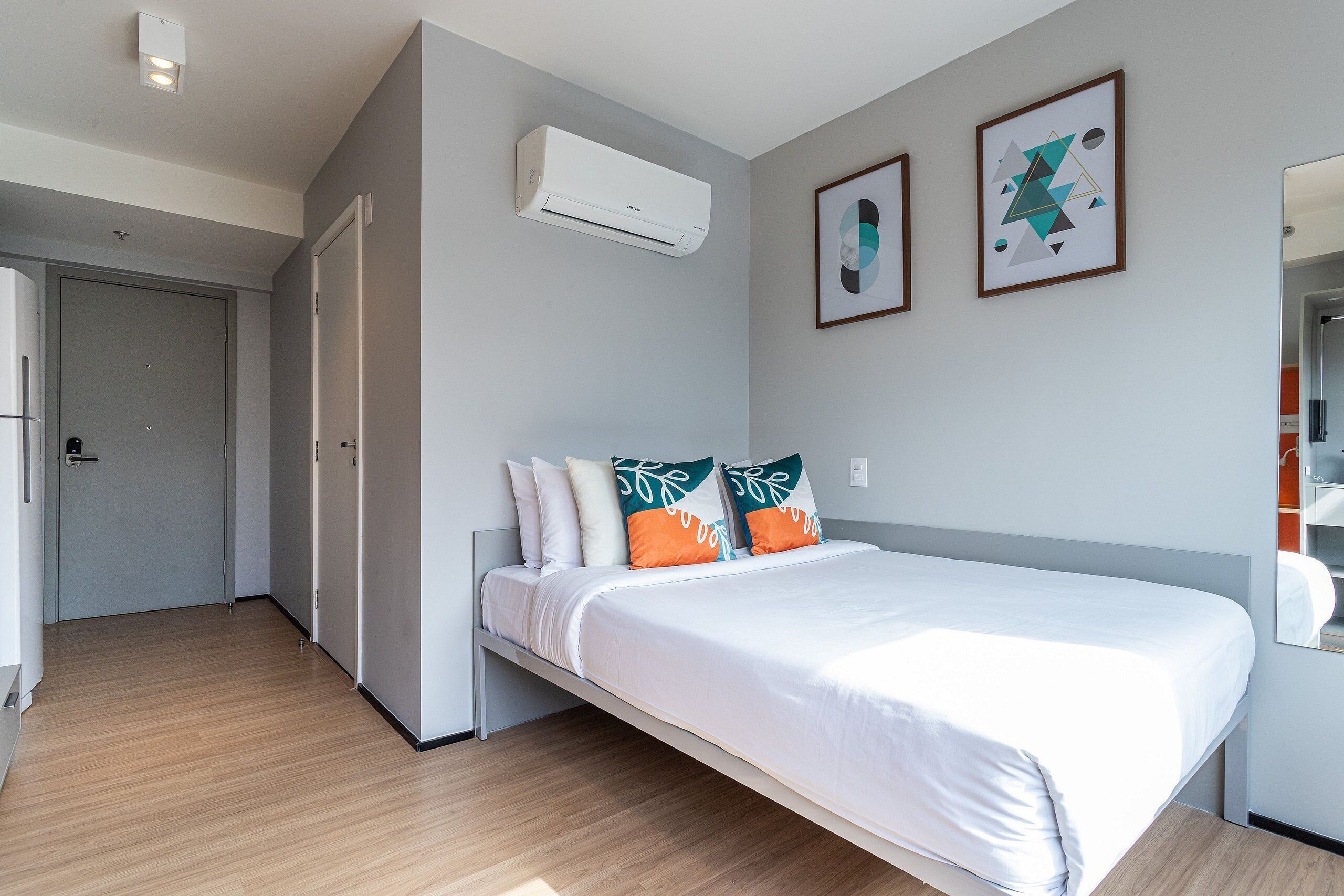 A modern hotel room with a bed, an air conditioning unit, a door, and a framed picture on the wall.