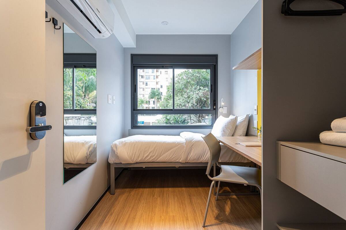 Ein kleines Hotelzimmer mit Bett, Schreibtisch, Stuhl, einem kleinen Badezimmer und großen Fenstern mit Blick auf Gebäude und Bäume.