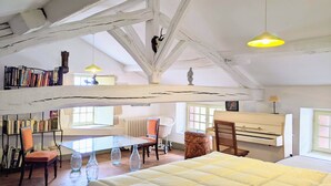 Un'ampia stanza mansardata con soffitto alto, travi a vista in legno, un letto grande, un pianoforte e una libreria piena di libri.