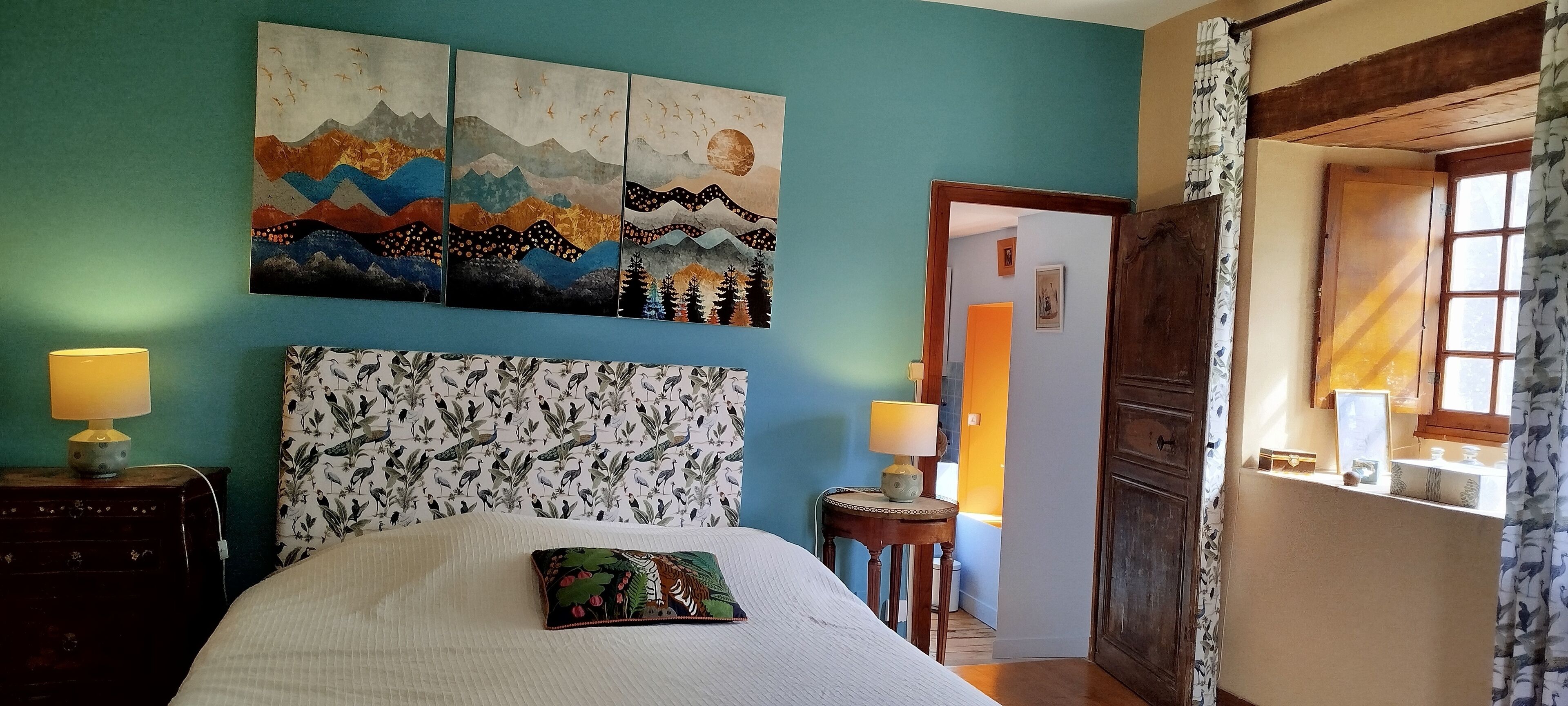 A bedroom with a large bed, two bedside tables with lamps, a wall with three paintings, and a door leading to another room.