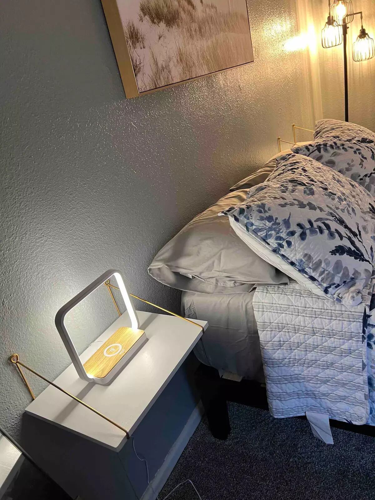 Une chambre à coucher avec une table de chevet, une lampe, un lit avec une literie à motifs floraux et un tableau accroché au mur.