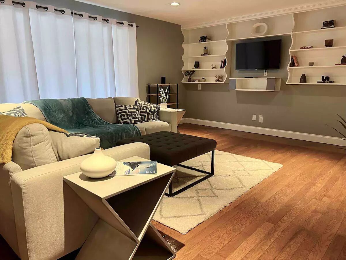 Un salon comprenant un canapé, un pouf et une table basse, un téléviseur à écran plat fixé au mur, ainsi que des étagères intégrées ornées d’objets décoratifs.