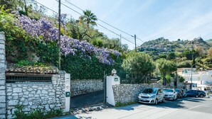 A stone wall with a gate, parked cars, and a hillside with purple flowers.