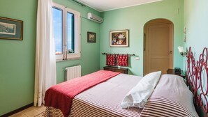 A bedroom with a bed, a window with curtains, a radiator, a door, and a wall-mounted air conditioner.