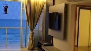 A modern hotel room with a large window offering a view of the sea, a flat-screen TV mounted on the wall, and a dining area with a table and chairs. - Apartamento Guarujá (Guarujá)