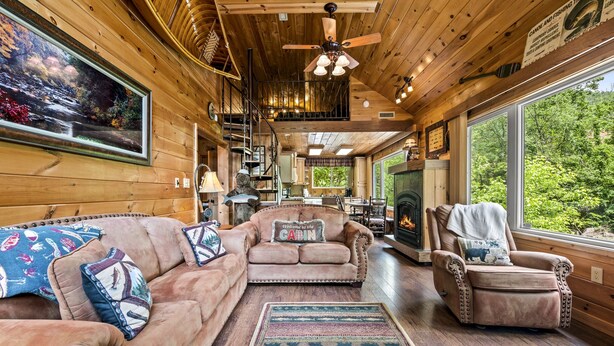 A cabin interior with a wooden interior, a fireplace, a sofa, a chair, and a large window overlooking greenery.