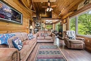 A cabin interior with a wooden interior, a fireplace, a sofa, a chair, and a large window overlooking greenery.
