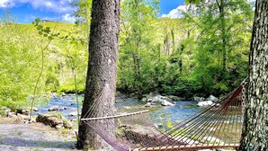 A hammock tied between trees by a river with a rocky bed and lush greenery in the background.