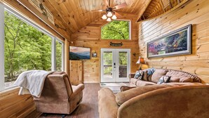 A cabin interior with wooden walls, a ceiling fan, a sofa, a chair, and a large window offering a view of trees.