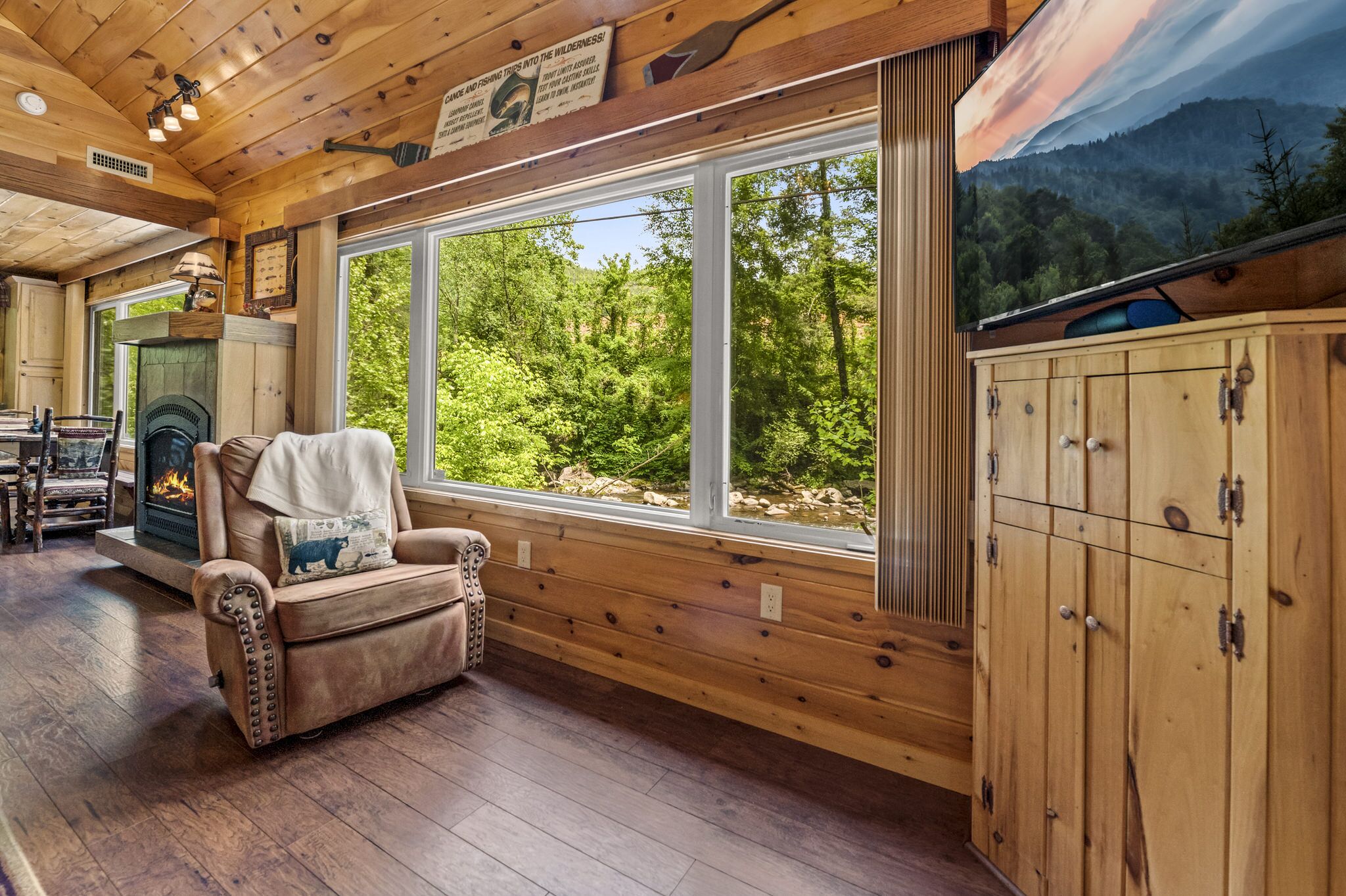 L’intérieur chaleureux d’un chalet avec une cheminée, un fauteuil en cuir et une grande fenêtre donnant sur une forêt.