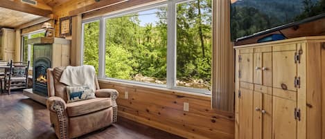 L’intérieur chaleureux d’un chalet avec une cheminée, un fauteuil en cuir et une grande fenêtre donnant sur une forêt.