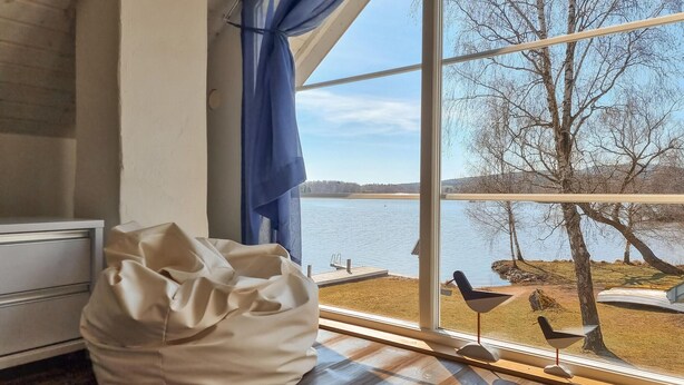 Une pièce dotée d’une grande fenêtre donnant sur un lac, d’un plancher en bois et d’une chaise blanche.