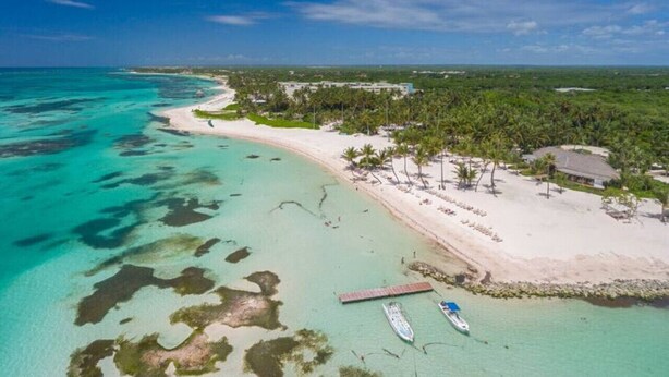 Luchtbeeld van een strand met helder turquoise water, een zandstrand en een kleine steiger met boten.