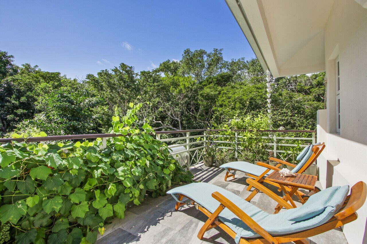 A balcony with two wooden lounge chairs and a table, surrounded by lush greenery and trees.