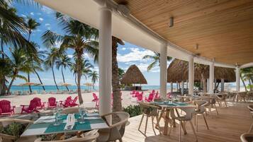 An outdoor dining area with tables and chairs, overlooking a beach with palm trees and a thatched-roof structure.
