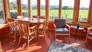 Un comedor con una mesa y sillas de madera, ventanas amplias con vistas a un campo verde, y una mesita auxiliar con una planta.
