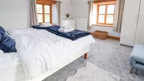 Un dormitorio con una cama grande, dos ventanas con cortinas, un radiador, una planta pequeña y una cesta de mimbre.